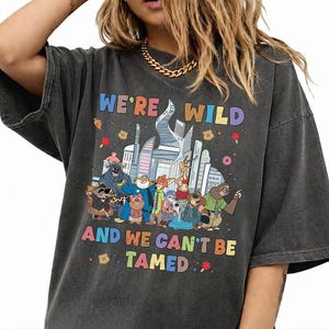 K&ouml;nnte beinhalten: Ein anthrazitfarbenes Oversize-T-Shirt mit einer bunten Grafik verschiedener animierter Tierfiguren vor einer Skyline. Der Text "WE'RE WILD" und "AND WE CAN'T BE TAMED" ist in mehrfarbigen Buchstaben oberhalb und unterhalb des Motivs gedruckt.