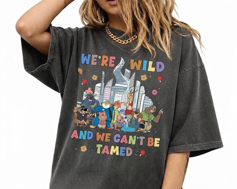K&ouml;nnte beinhalten: Ein anthrazitfarbenes Oversize-T-Shirt mit einer bunten Grafik verschiedener animierter Tierfiguren vor einer Skyline. Der Text "WE'RE WILD" und "AND WE CAN'T BE TAMED" ist in mehrfarbigen Buchstaben oberhalb und unterhalb des Motivs gedruckt.