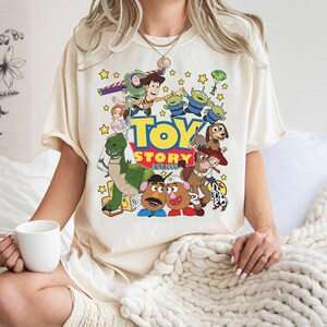 May include: Cream-colored t-shirt featuring a colorful graphic of characters from the animated film "Toy Story." The design includes Woody, Buzz Lightyear, and other characters, with the words "TOY STORY EST 1995" in a playful font. The shirt has short sleeves.