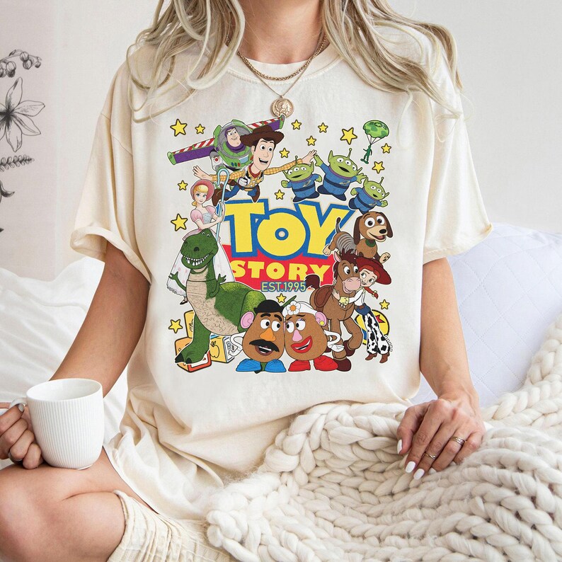 May include: Cream-colored t-shirt featuring a colorful graphic of characters from the animated film "Toy Story." The design includes Woody, Buzz Lightyear, and other characters, with the words "TOY STORY EST 1995" in a playful font. The shirt has short sleeves.