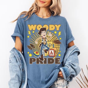 May include: Blue t-shirt featuring a graphic of Woody from Toy Story. The design includes the word "WOODY" above the character and "PRIDE" below. Additional elements include stars, a hat, and a toy block.