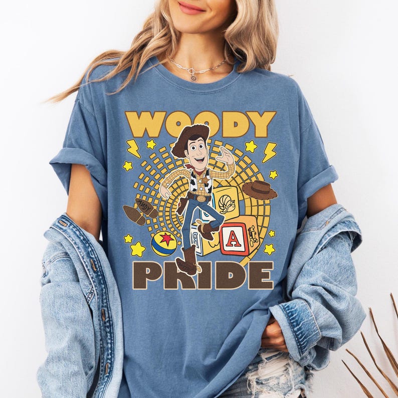 May include: Blue t-shirt featuring a graphic of Woody from Toy Story. The design includes the word "WOODY" above the character and "PRIDE" below. Additional elements include stars, a hat, and a toy block.