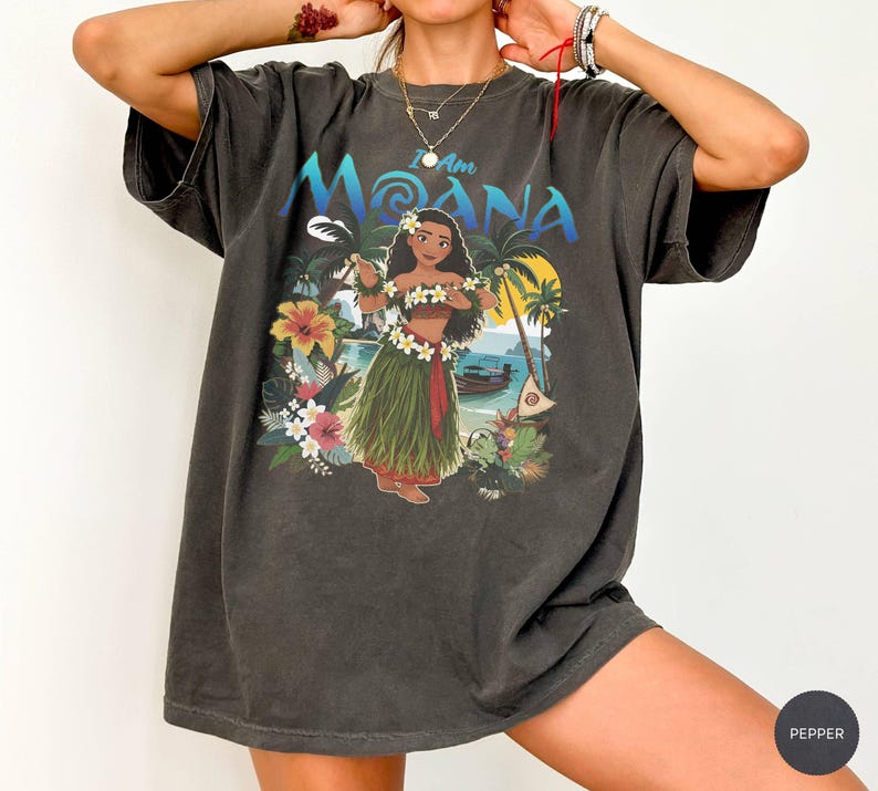 May include: Dark gray t-shirt featuring the phrase "I Am Moana" above a graphic of Moana in a floral lei, standing in front of a tropical scene with palm trees, a boat, and flowers. The shirt has a vintage, worn look.