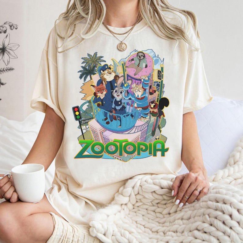Puede incluir: Camiseta color crema con un gr&aacute;fico colorido de la pel&iacute;cula animada "Zootopia". El dise&ntilde;o incluye varios personajes de animales y el t&iacute;tulo de la pel&iacute;cula en verde y amarillo. La camiseta la lleva una persona.