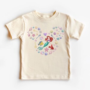 May include: Cream-colored t-shirt featuring a heart-shaped design composed of seashells, starfish, and a mermaid. The mermaid has red hair and a green tail. Other sea creatures and utensils are also included in the design.