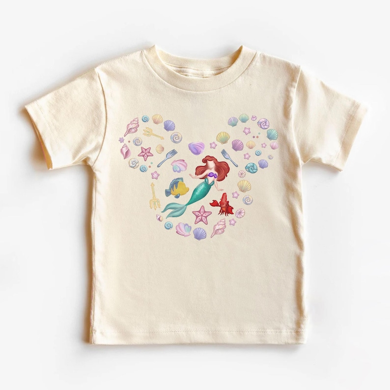 May include: Cream-colored t-shirt featuring a heart-shaped design composed of seashells, starfish, and a mermaid. The mermaid has red hair and a green tail. Other sea creatures and utensils are also included in the design.
