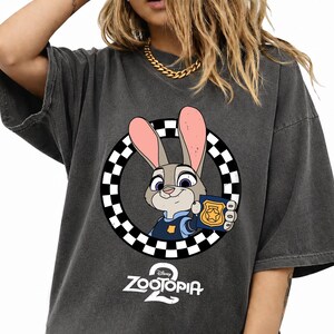 May include: A charcoal gray short-sleeve t-shirt featuring a graphic of a cartoon rabbit in a blue uniform holding a gold police badge inside a black and white checkered circle, with the text Zootopia 2 printed in white below the graphic.