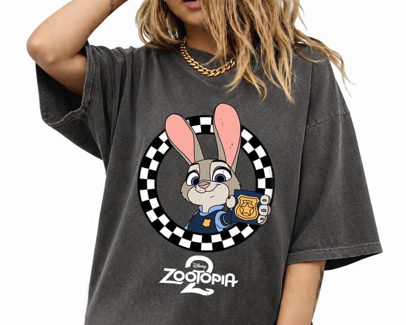 May include: A charcoal gray short-sleeve t-shirt featuring a graphic of a cartoon rabbit in a blue uniform holding a gold police badge inside a black and white checkered circle, with the text Zootopia 2 printed in white below the graphic.