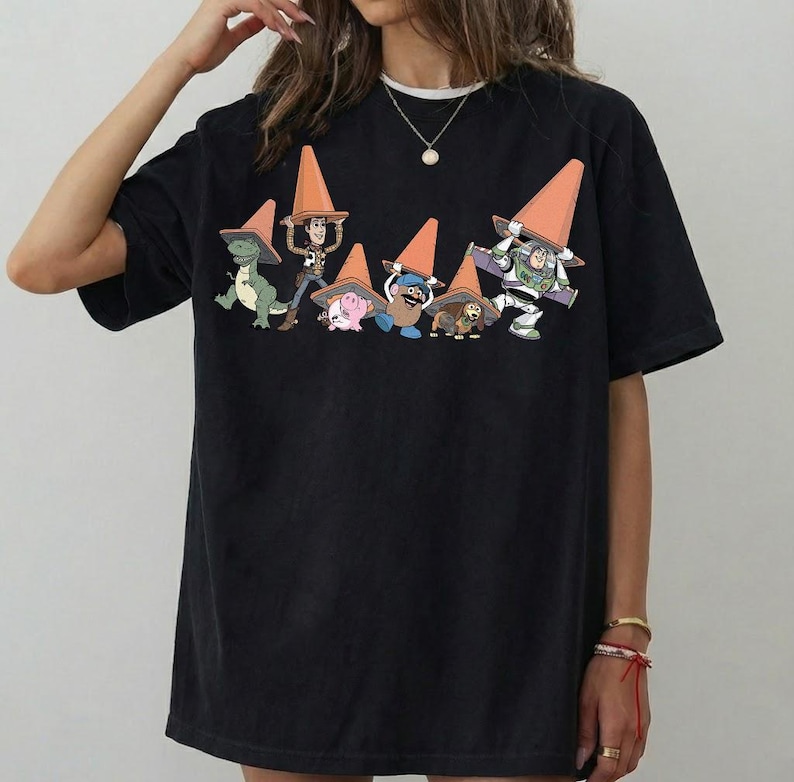 May include: Black t-shirt featuring a graphic of Toy Story characters wearing orange traffic cones. The characters include Rex, Woody, Hamm, Mr. Potato Head, Slinky Dog, and Buzz Lightyear. The shirt has a crew neck and short sleeves.