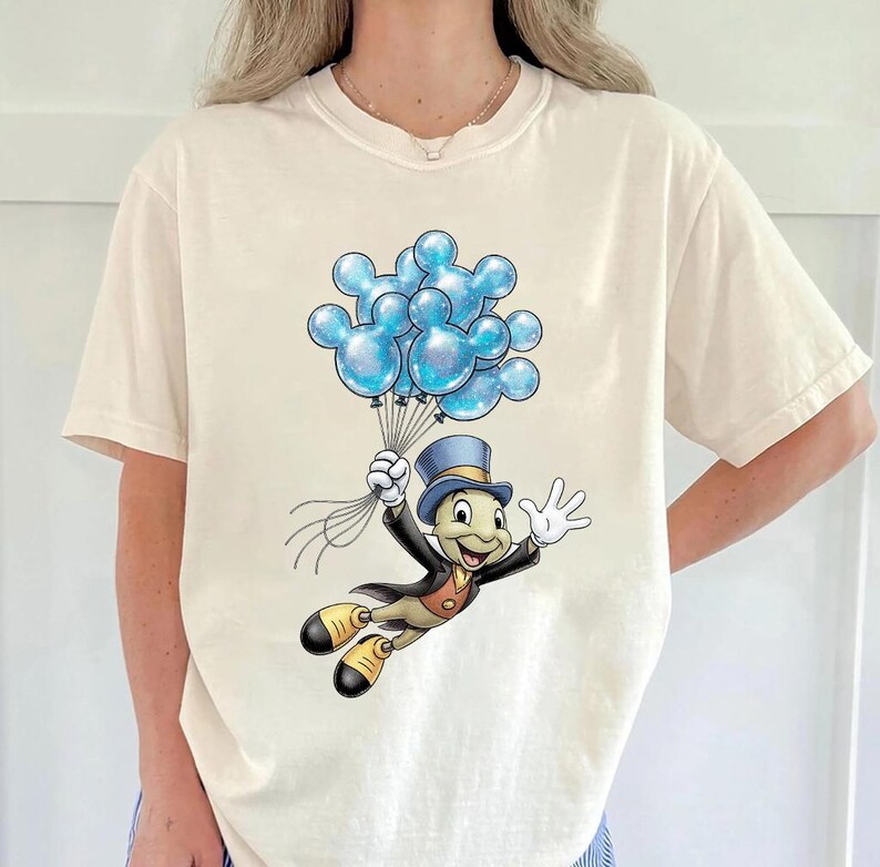 May include: Cream-colored t-shirt featuring a cartoon illustration of Jiminy Cricket holding a bunch of blue Mickey Mouse-shaped balloons. Jiminy Cricket is wearing a top hat, black suit, and yellow shoes. The balloons have a glittery effect.
