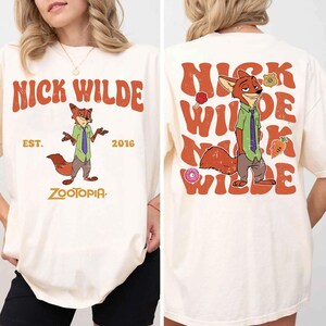 Pode incluir: Uma t-shirt em tom creme com um gr&aacute;fico da personagem Nick Wilde de Zootopia. A frente exibe a personagem com o texto NICK WILDE, EST. 2016 e ZOOTOPIA. As costas apresentam um gr&aacute;fico grande da personagem com texto repetido.