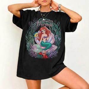 May include: Black t-shirt featuring a colorful graphic of Ariel from The Little Mermaid, seated in a seashell. The design includes Flounder, Sebastian, and aquatic plants. The text "The Little Mermaid" is arched above the image.