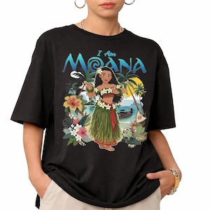 May include: Black t-shirt featuring a colorful graphic of a Polynesian woman with the text "I Am Moana". The design includes tropical flowers, palm trees, and a boat on the water. The shirt is a crew neck.