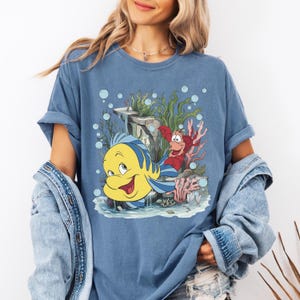 Puede incluir: Camiseta azul con un dibujo animado de Flounder y Sebasti&aacute;n de La Sirenita. El dise&ntilde;o incluye elementos submarinos como coral, algas y burbujas. La camiseta es de color azul liso.