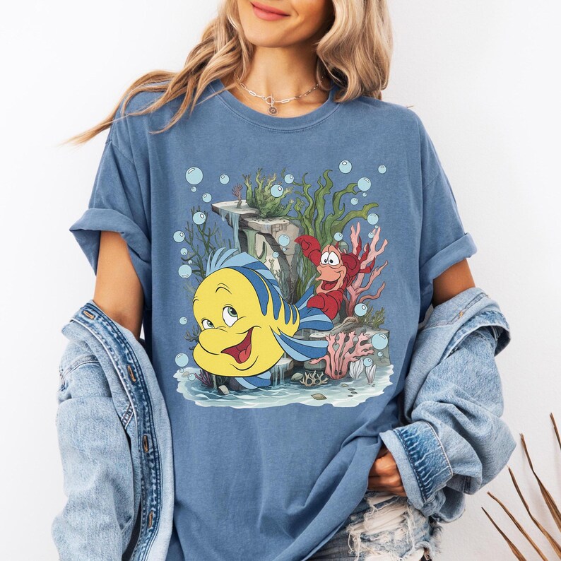 Puede incluir: Camiseta azul con un dibujo animado de Flounder y Sebasti&aacute;n de La Sirenita. El dise&ntilde;o incluye elementos submarinos como coral, algas y burbujas. La camiseta es de color azul liso.
