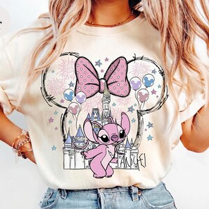 Puede incluir: Una camiseta color marfil con un estampado gr&aacute;fico de un personaje alien&iacute;gena rosa frente al contorno de un castillo. El dise&ntilde;o incluye un lazo grande de lunares, fuegos artificiales y globos dispuestos en una silueta de orejas de rat&oacute;n.
