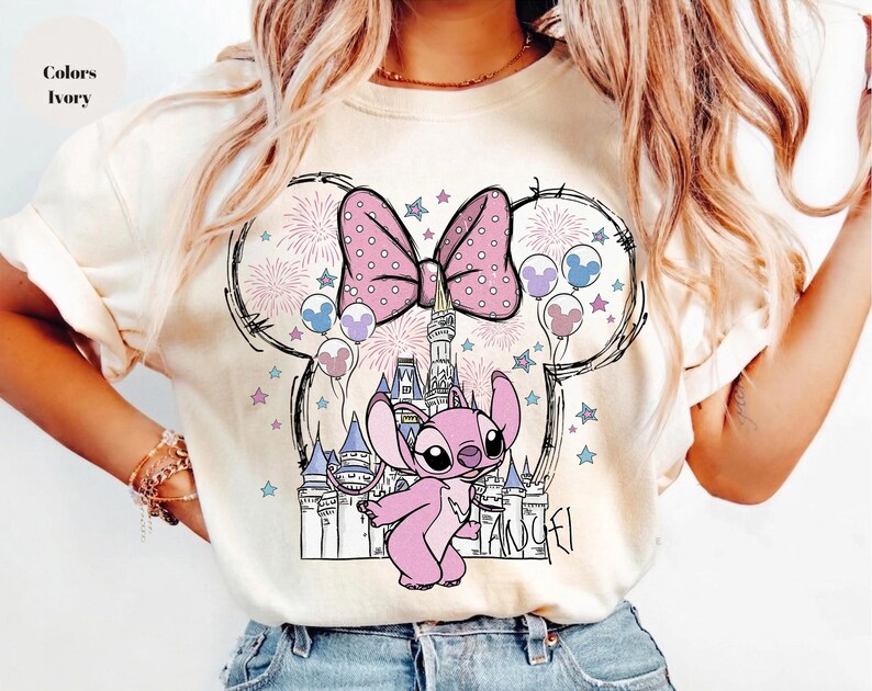 Puede incluir: Una camiseta color marfil con un estampado gr&aacute;fico de un personaje alien&iacute;gena rosa frente al contorno de un castillo. El dise&ntilde;o incluye un lazo grande de lunares, fuegos artificiales y globos dispuestos en una silueta de orejas de rat&oacute;n.