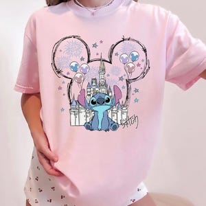 Puede incluir: Una camiseta de color rosa claro con un gr&aacute;fico del personaje Stitch sentado frente al contorno de un castillo. El dise&ntilde;o incluye una silueta de orejas de Mickey Mouse rellena de fuegos artificiales, estrellas y globos, con el nombre Stitch en negro.
