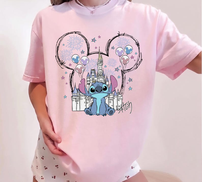 Puede incluir: Una camiseta de color rosa claro con un gr&aacute;fico del personaje Stitch sentado frente al contorno de un castillo. El dise&ntilde;o incluye una silueta de orejas de Mickey Mouse rellena de fuegos artificiales, estrellas y globos, con el nombre Stitch en negro.