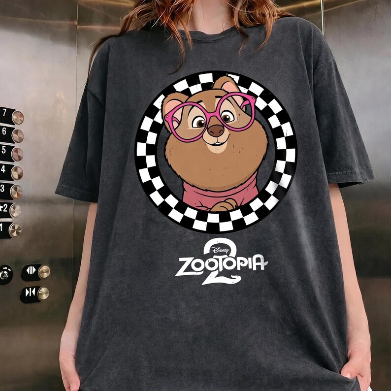 May include: A charcoal gray short-sleeve t-shirt featuring a graphic of a brown animal wearing pink glasses inside a black and white checkered circle. Below the circle, the white text reads Disney Zootopia 2 in a stylized font.