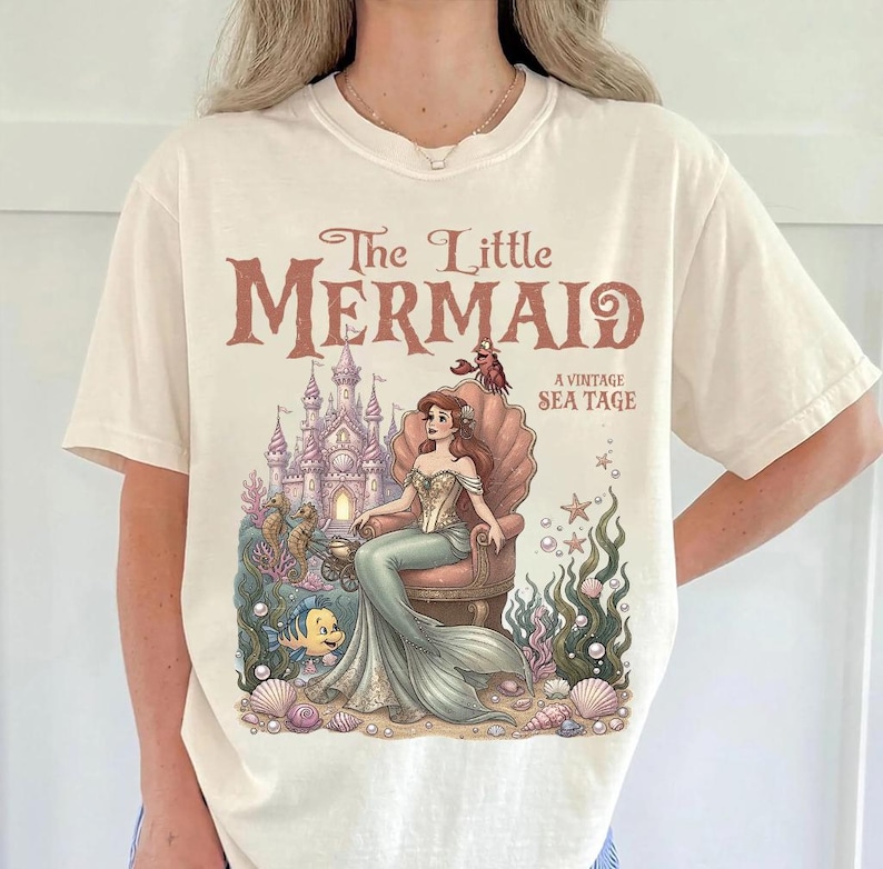 May include: Cream-colored t-shirt featuring a vintage-style illustration from "The Little Mermaid." The design includes the title, a mermaid seated on a shell throne, a castle, and sea creatures. The shirt has short sleeves and a crew neck.