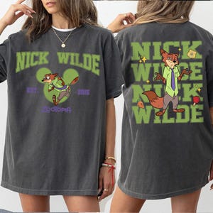 May include: A charcoal gray t-shirt featuring Nick Wilde from Zootopia. The front displays the text NICK WILDE, a green Mickey Mouse silhouette, and the text EST. 2016 ZOOTOPIA. The back shows a large graphic of Nick Wilde with the text NICK WILDE.