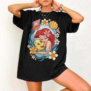 May include: Black t-shirt featuring a colorful graphic of Ariel from The Little Mermaid hugging Flounder. The design is framed by seashells, flowers, and ocean waves. The shirt has short sleeves and a relaxed fit.