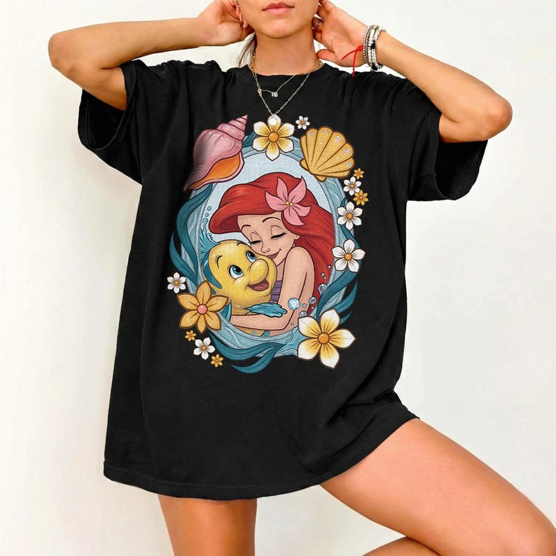 May include: Black t-shirt featuring a colorful graphic of Ariel from The Little Mermaid hugging Flounder. The design is framed by seashells, flowers, and ocean waves. The shirt has short sleeves and a relaxed fit.