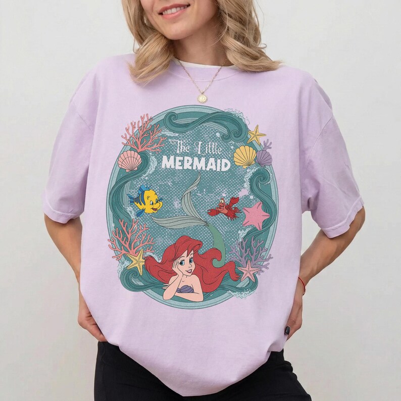 May include: A light purple t-shirt featuring a circular graphic from "The Little Mermaid." The graphic includes Ariel, Flounder, Sebastian, and the text "The Little Mermaid" in a teal circle with coral and seashells.