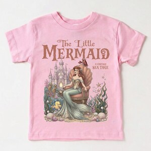 May include: Pink t-shirt featuring a vintage-style illustration of The Little Mermaid. The design includes Ariel seated on a seashell throne, a castle, and sea creatures. Text reads "The Little Mermaid A Vintage Sea Tage."