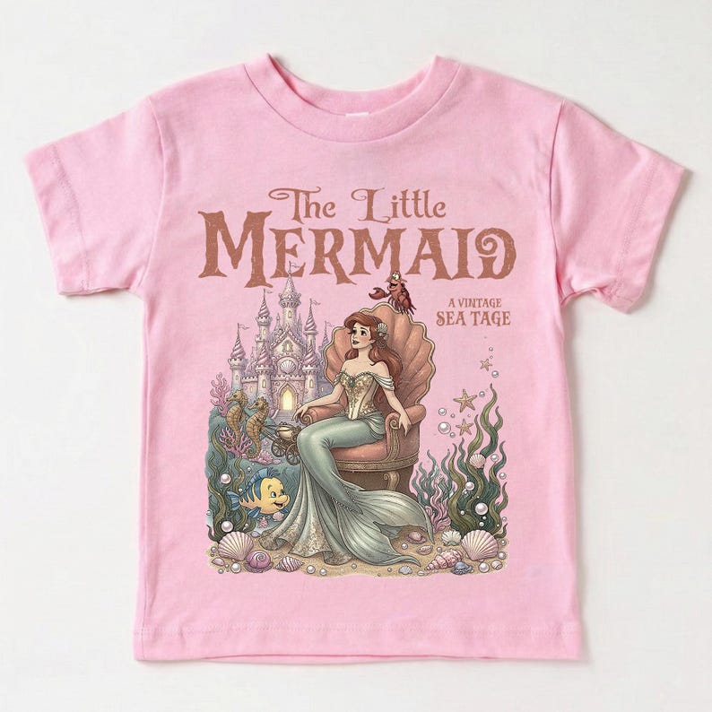 May include: Pink t-shirt featuring a vintage-style illustration of The Little Mermaid. The design includes Ariel seated on a seashell throne, a castle, and sea creatures. Text reads "The Little Mermaid A Vintage Sea Tage."