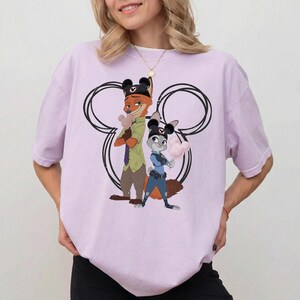 May include: A light purple short-sleeve t-shirt featuring a graphic of Nick Wilde and Judy Hopps from Zootopia, both wearing black mouse ear hats. They are positioned in front of a large black circular outline resembling a mouse head silhouette.