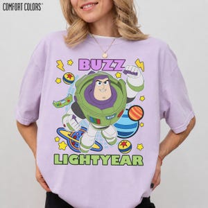 May include: Lilac Comfort Colors t-shirt featuring a cartoon graphic of Buzz Lightyear. The design includes the word "BUZZ" in purple and "LIGHTYEAR" in green, along with planets, stars, and lightning bolts. The shirt is a relaxed fit.