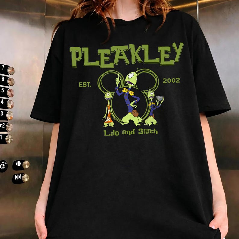 Puede incluir: Una camiseta negra de manga corta con un estampado gr&aacute;fico del personaje Pleakley de Lilo y Stitch. El texto dice PLEAKLEY EST. 2002 y Lilo and Stitch, con tres figuras alien&iacute;genas verdes frente a un contorno circular.