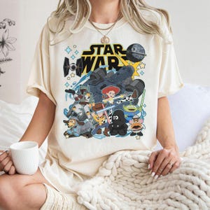 May include: Cream-colored t-shirt featuring a graphic of Star Wars characters and the Star Wars logo. The design includes characters from both Star Wars and Toy Story, with a blue and yellow color scheme. The shirt is short-sleeved.