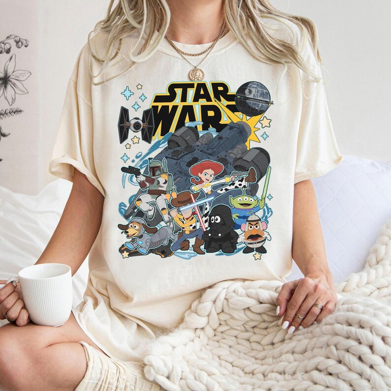 May include: Cream-colored t-shirt featuring a graphic of Star Wars characters and the Star Wars logo. The design includes characters from both Star Wars and Toy Story, with a blue and yellow color scheme. The shirt is short-sleeved.