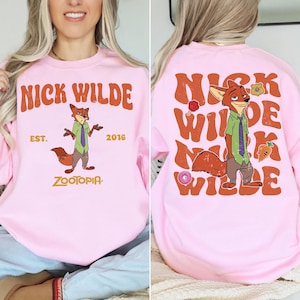 Pode incluir: Uma camisola de gola redonda rosa claro com um gr&aacute;fico da personagem Nick Wilde de Zootopia. A frente exibe o texto NICK WILDE EST. 2016 ZOOTOPIA. As costas apresentam um grande texto NICK WILDE repetido com a personagem.