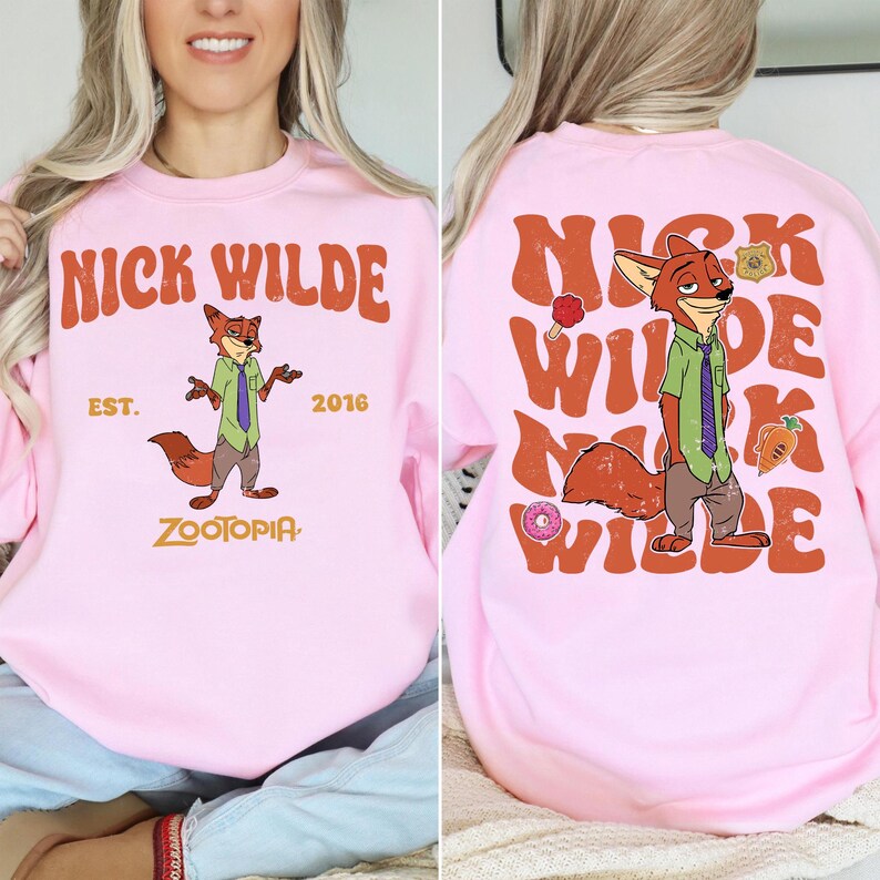 Pode incluir: Uma camisola de gola redonda rosa claro com um gr&aacute;fico da personagem Nick Wilde de Zootopia. A frente exibe o texto NICK WILDE EST. 2016 ZOOTOPIA. As costas apresentam um grande texto NICK WILDE repetido com a personagem.