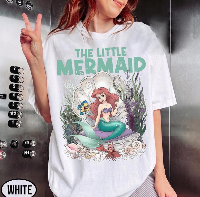 May include: White t-shirt featuring a graphic of Ariel from The Little Mermaid. The design includes Ariel sitting in a seashell, with Flounder and Sebastian. The text "THE LITTLE MERMAID" is printed above the image in a teal color.