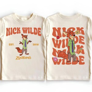 Pode incluir: Uma camiseta cor de creme com Nick Wilde de Zootopia. A frente exibe o personagem com o texto NICK WILDE EST. 2016 ZOOTOPIA. As costas apresentam o personagem repetido com o texto NICK WILDE, um donut e uma cenoura.