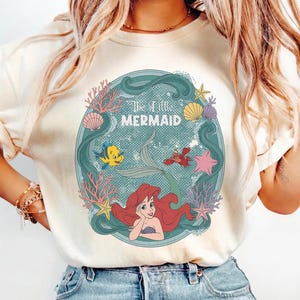 May include: Cream-colored t-shirt featuring a circular graphic from "The Little Mermaid." The design includes Ariel, Flounder, Sebastian, and other sea elements. The words "The Little Mermaid" are at the top of the graphic.