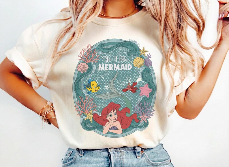 May include: Cream-colored t-shirt featuring a circular graphic from "The Little Mermaid." The design includes Ariel, Flounder, Sebastian, and other sea elements. The words "The Little Mermaid" are at the top of the graphic.