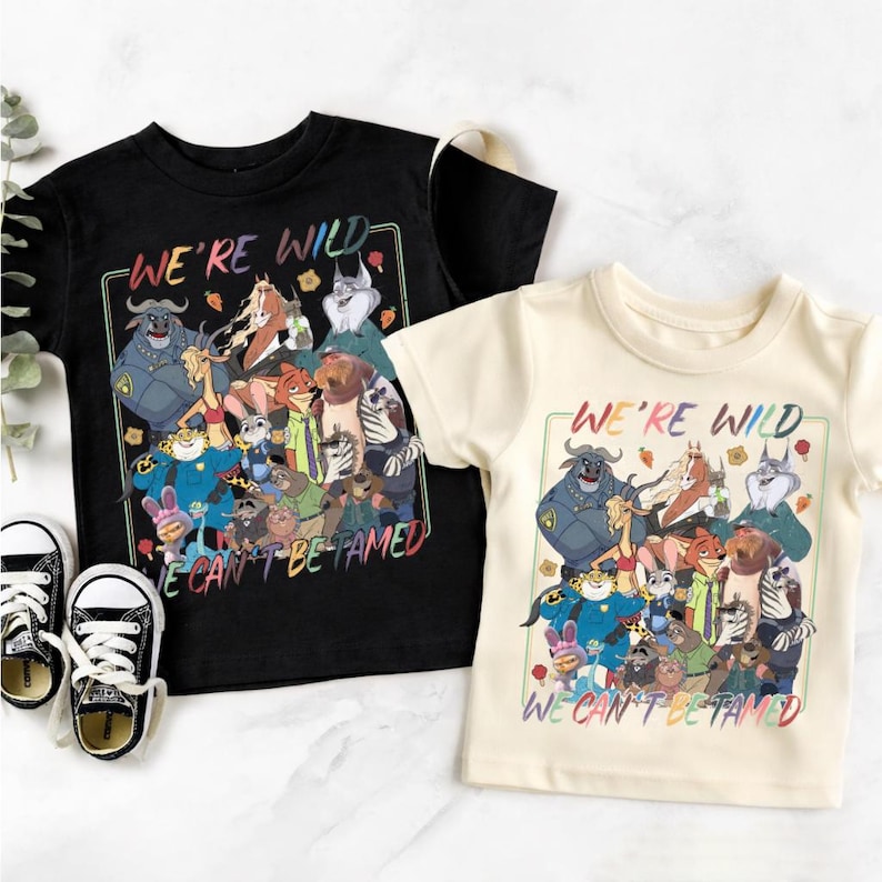 May include: A black t-shirt and a cream t-shirt, each featuring a colorful graphic print of various animated animal characters. The text "WE'RE WILD" appears at the top and "WE CAN'T BE TAMED" at the bottom of the rectangular graphic design.