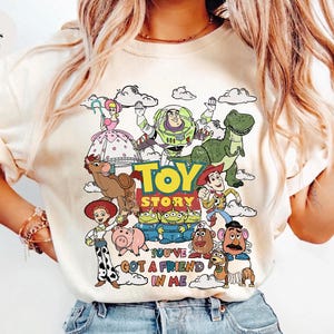 Op de afbeelding: Ivoor t-shirt met een kleurrijke Toy Story-afbeelding, met Woody, Buzz Lightyear en Rex. De tekst "TOY STORY" en "YOU'VE GOT A FRIEND IN ME" zijn ook te zien. Een leuk, geanimeerd ontwerp.