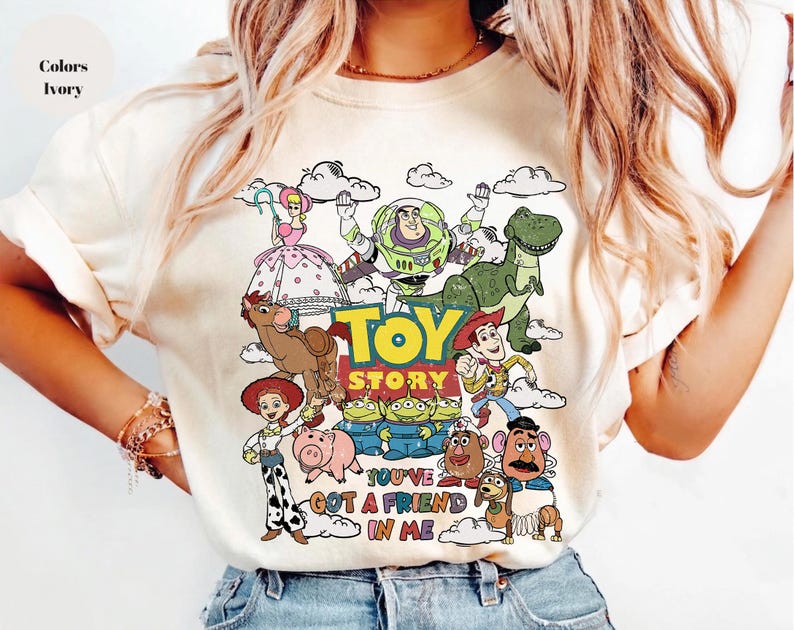Op de afbeelding: Ivoor t-shirt met een kleurrijke Toy Story-afbeelding, met Woody, Buzz Lightyear en Rex. De tekst "TOY STORY" en "YOU'VE GOT A FRIEND IN ME" zijn ook te zien. Een leuk, geanimeerd ontwerp.