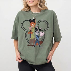 May include: An olive green short-sleeve t-shirt featuring a graphic print of two animated fox and rabbit characters wearing black mouse-ear hats. The characters are positioned in front of a large black outline of a mouse head silhouette.