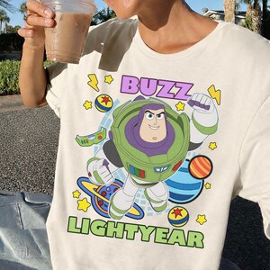 May include: Cream-colored t-shirt featuring a graphic of Buzz Lightyear from Toy Story. The design includes the words "BUZZ LIGHTYEAR" in green and purple, with stars and planets. The shirt is a casual wear item.