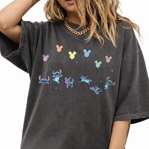May include: A charcoal gray oversized t-shirt featuring a graphic print of the character Stitch from Lilo and Stitch. The design shows multiple versions of the blue character floating while holding onto colorful pastel Mickey Mouse shaped balloons.