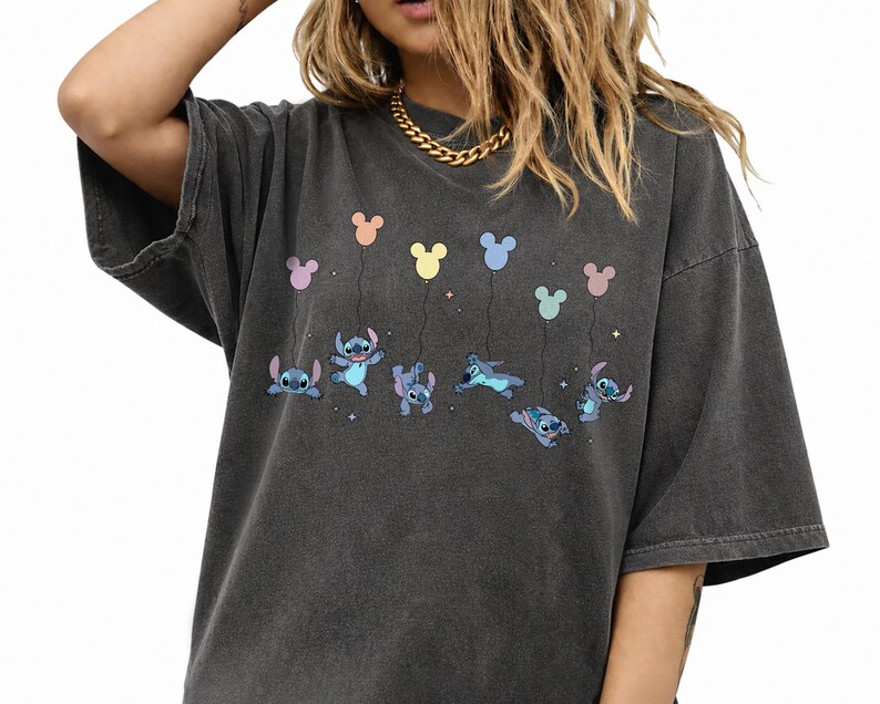 May include: A charcoal gray oversized t-shirt featuring a graphic print of the character Stitch from Lilo and Stitch. The design shows multiple versions of the blue character floating while holding onto colorful pastel Mickey Mouse shaped balloons.