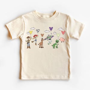 Op de afbeelding: Cr&egrave;mekleurig T-shirt met een kleurrijke afbeelding van stripfiguren die ballonvormige ballonnen vasthouden. De personages zijn onder meer een cowboy, cowgirl, paard, ruimteranger, dinosaurus en varken. De ballonnen zijn in verschillende kleuren en vormen.